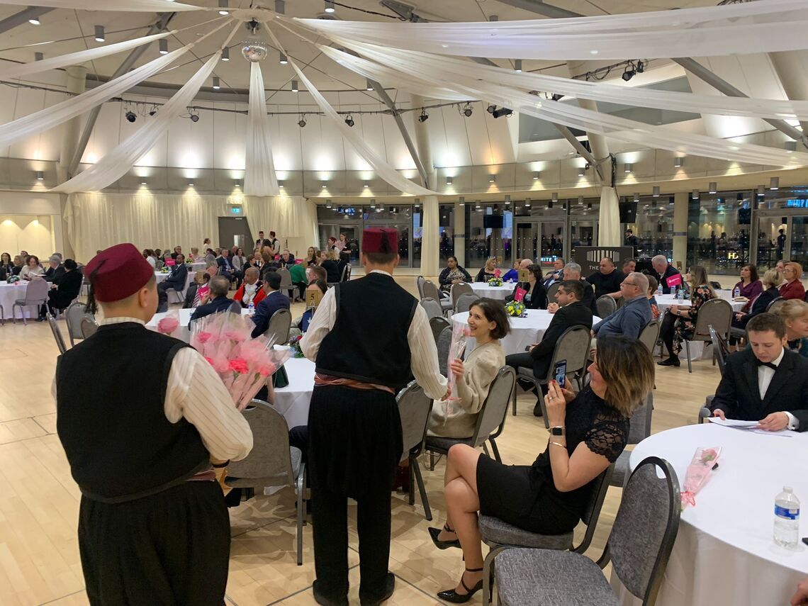 People at a large cultural event sit at tables smiling and chatting, with two men in traditional Bosnian men's costumes facing the crowd